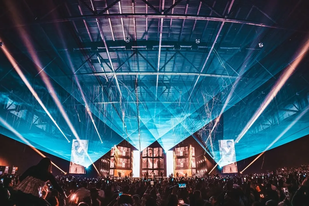 Arena filled with converging teal beams above the LED panels, crowd silhouetted in the foreground