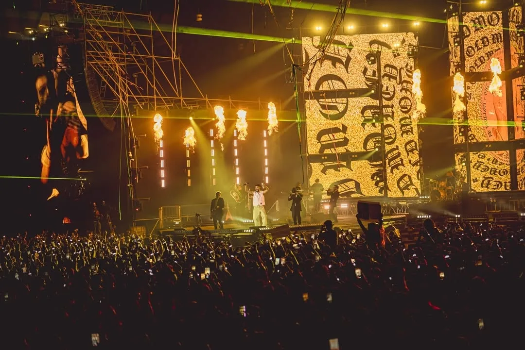 Outdoor night stage, gold lettering artwork on the screens above pyro jets and a crowd of phones