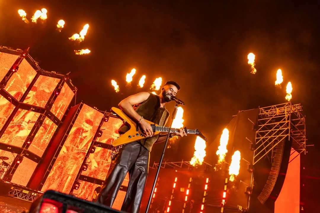 The artist with a guitar between tall flame jets, warm artwork on the panels behind