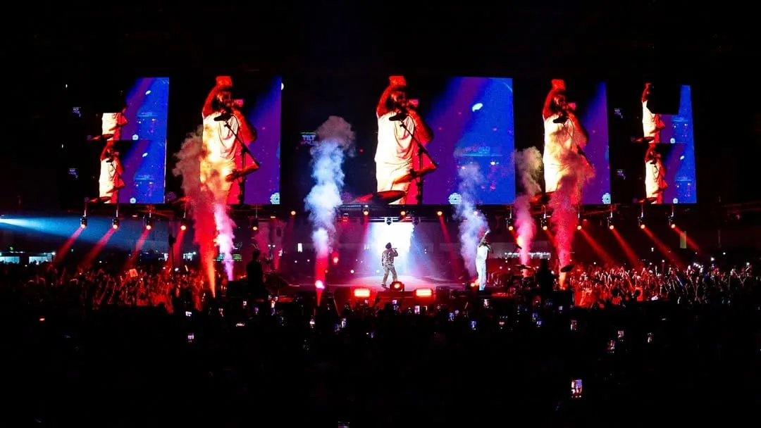 Dark venue with a row of panels showing figures, red and white beams cutting through smoke